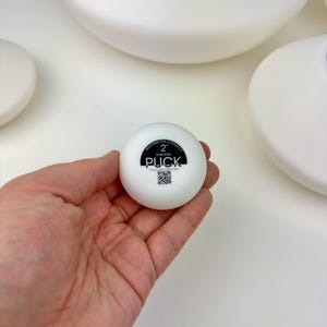 May include: A small, round, white ceramic object with a black circular label on the top. The label features the text MOONMOON CERAMICS, 2, SHROOM, PUCK, and a small QR code. The object has a smooth, matte finish and a compact, circular shape.