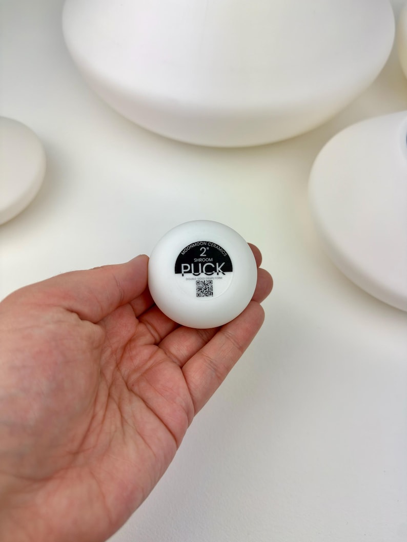 May include: A small, round, white ceramic object with a black circular label on the top. The label features the text MOONMOON CERAMICS, 2, SHROOM, PUCK, and a small QR code. The object has a smooth, matte finish and a compact, circular shape.