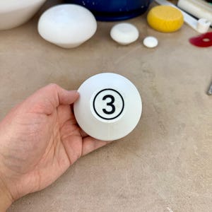 May include: A white, bowl-shaped ceramic object with a circular black outline containing the number 3 printed in the center. The object has a smooth, matte finish and a rounded, tapered base, displaying a clear numerical marking on its surface.
