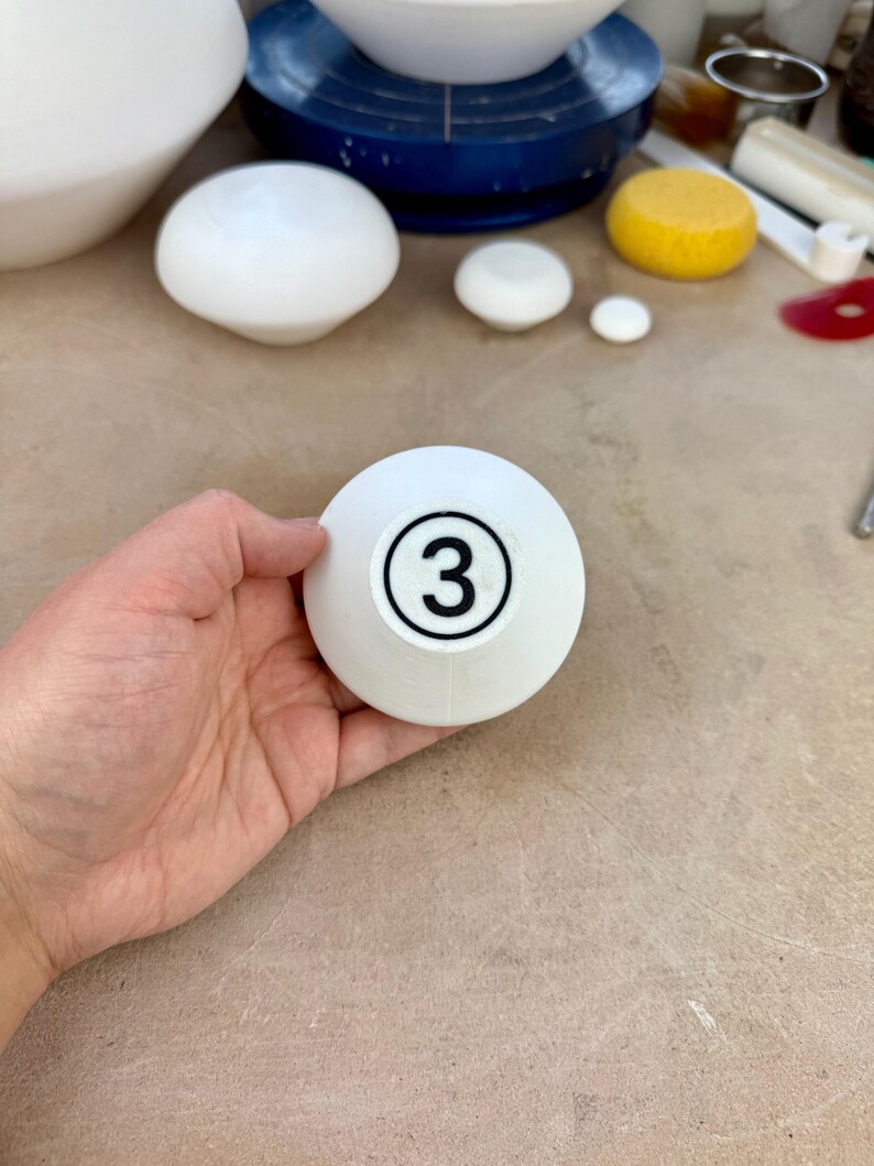 May include: A white, bowl-shaped ceramic object with a circular black outline containing the number 3 printed in the center. The object has a smooth, matte finish and a rounded, tapered base, displaying a clear numerical marking on its surface.