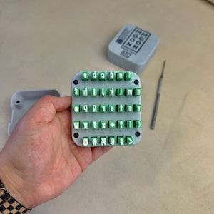 May include: A square gray stamp block featuring a grid of raised green rubber letters, numbers, and symbols. The set includes a matching gray storage case with the text "MOON ZOO" and a thin metal stylus tool for arranging the characters.