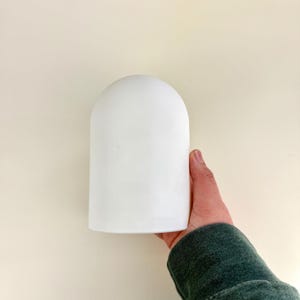 Op de afbeelding: Een wit, cilindrisch object met een afgeronde bovenkant, vastgehouden door een persoon. Het object is glad en ondoorzichtig, met een eenvoudig, minimalistisch ontwerp. De achtergrond is effen lichtbeige.