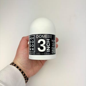 Puede incluir: Un molde blanco de cerámica en forma de cúpula, de aproximadamente 7,6 cm de diámetro, sostenido en una mano. El molde tiene una etiqueta en blanco y negro con el texto "DOME DRAPE MOLD 3 INCH" y "MM OO NN CERAMICS made in Detroit, Michigan". También se ve un código QR.