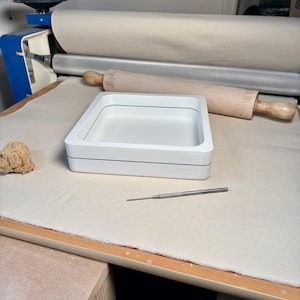 May include: A square white tray with a thin black line detail along the side, positioned on a flat surface. The tray features a smooth, matte finish and a hollow center, accompanied by a small metal tool and a wooden rolling pin nearby.