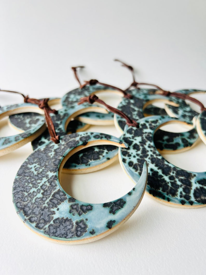 May include: A group of ceramic crescent moon ornaments with a blue and black speckled glaze. The ornaments have brown leather cords attached to them.
