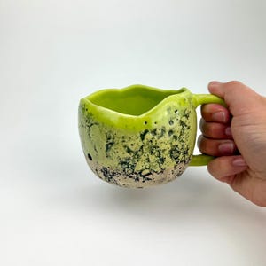 Op de afbeelding: Een handgemaakte keramische mok met een uniek, getextureerd oppervlak. De mok heeft een felgroen interieur en handvat, met een gespikkelde, lichtgroene en zwarte buitenkant. De mok wordt door een hand tegen een witte achtergrond gehouden.