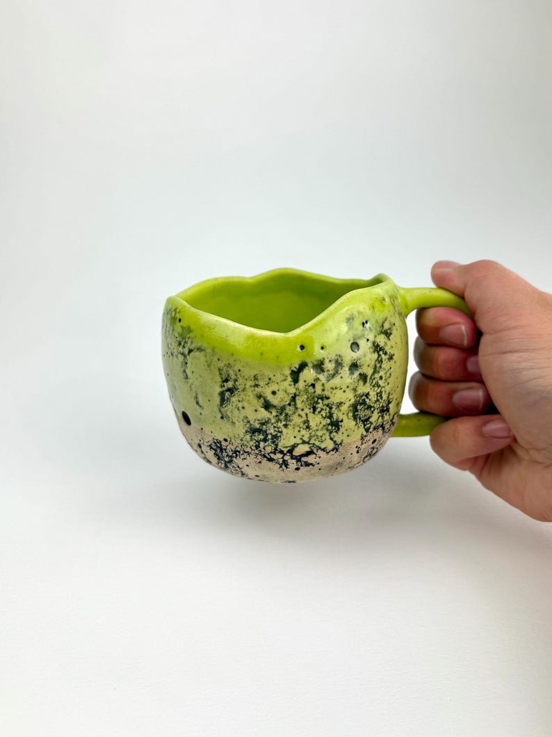 Op de afbeelding: Een handgemaakte keramische mok met een uniek, getextureerd oppervlak. De mok heeft een felgroen interieur en handvat, met een gespikkelde, lichtgroene en zwarte buitenkant. De mok wordt door een hand tegen een witte achtergrond gehouden.