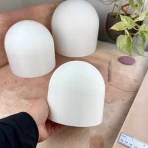 May include: Three white, smooth, dome-shaped objects with a cylindrical base. One dome is held in the foreground, while two others sit behind it. A metal ruler is visible in the corner, showing measurements in inches for scale reference.