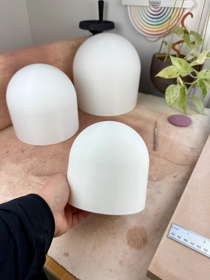May include: Three white, smooth, dome-shaped objects with a cylindrical base. One dome is held in the foreground, while two others sit behind it. A metal ruler is visible in the corner, showing measurements in inches for scale reference.