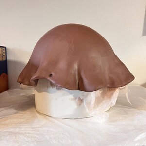 May include: A smooth, brown, dome-shaped clay form with a wide, flared, wavy edge draped over a cylindrical white support structure. The clay surface is uniform in color and texture, featuring a small, circular hole near the lower edge.