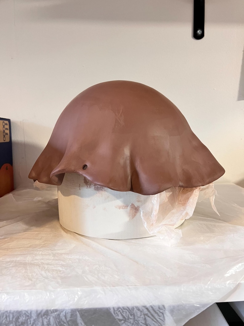 May include: A smooth, brown, dome-shaped clay form with a wide, flared, wavy edge draped over a cylindrical white support structure. The clay surface is uniform in color and texture, featuring a small, circular hole near the lower edge.