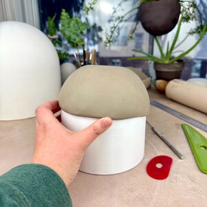 May include: A cylindrical white ceramic vessel topped with a smooth, rounded, light gray clay dome. The object features a clean, matte finish on the white base and a soft, unglazed texture on the gray top section, creating a two-tone appearance.