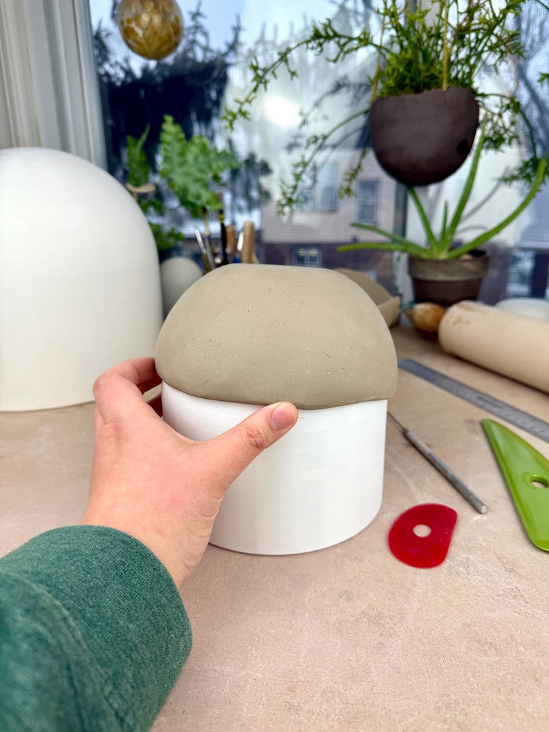 May include: A cylindrical white ceramic vessel topped with a smooth, rounded, light gray clay dome. The object features a clean, matte finish on the white base and a soft, unglazed texture on the gray top section, creating a two-tone appearance.