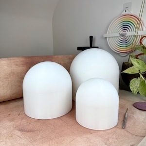 May include: Three white, smooth, dome-shaped objects of varying sizes arranged in a cluster. Each piece features a cylindrical base that transitions into a rounded top, creating a uniform, matte appearance across all three distinct sculptural forms.
