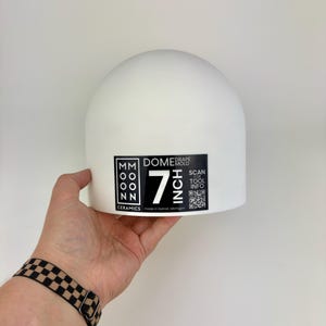 May include: A white ceramic dome drape mold with a black label featuring the text MOON MOON CERAMICS, DOME DRAPE MOLD, 7 INCH, and SCAN FOR TOOL INFO with a QR code. The mold is a smooth, semi-spherical object designed for pottery and clay forming.