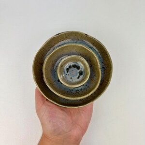 May include: Three nested ceramic bowls with a circular shape, featuring a speckled olive-green exterior and a smooth, glossy interior glaze that transitions from a deep blue center to a lighter, mottled brown rim with dark brown speckles.
