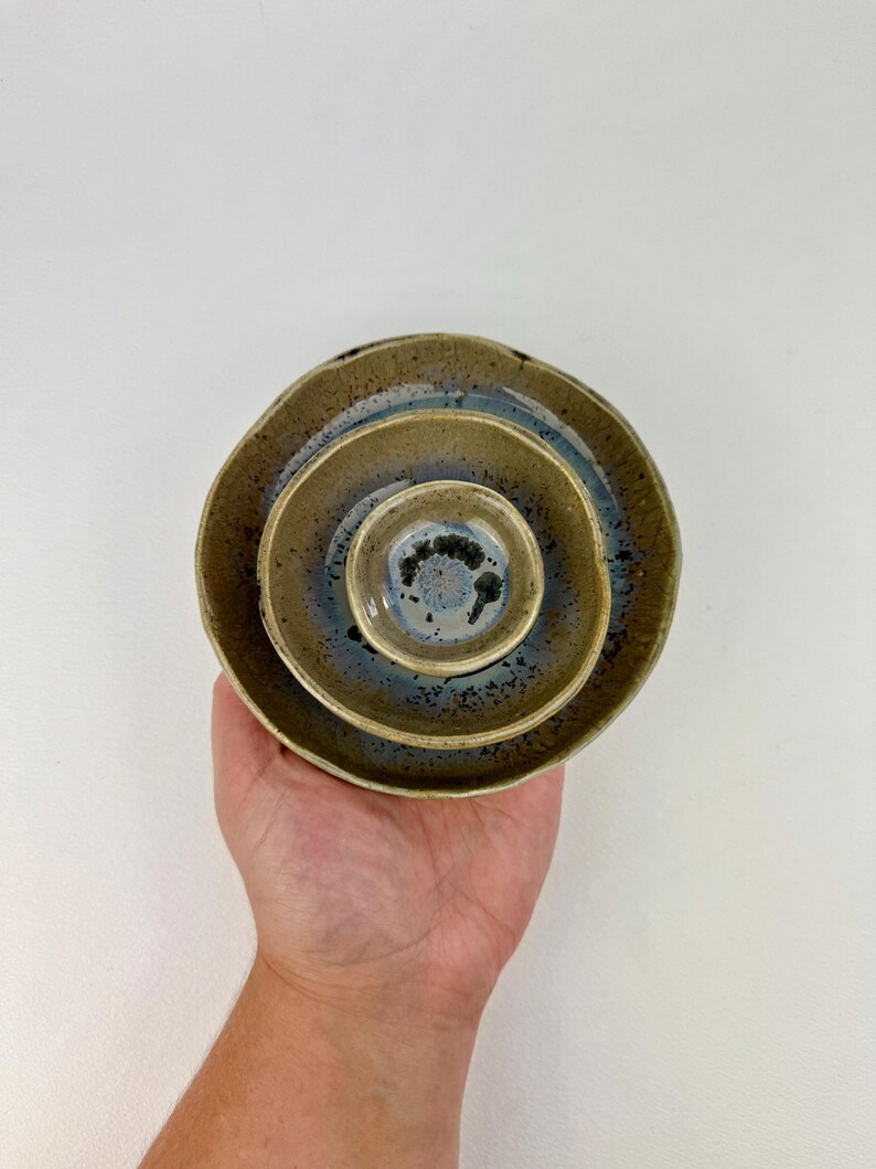 May include: Three nested ceramic bowls with a circular shape, featuring a speckled olive-green exterior and a smooth, glossy interior glaze that transitions from a deep blue center to a lighter, mottled brown rim with dark brown speckles.