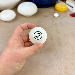 May include: A smooth white spherical object featuring a black circular outline with the number 2 printed in the center. The object is held in a position that displays the clear black numeral against the solid white surface of the sphere.