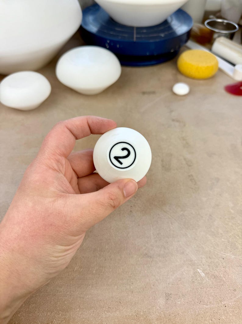 May include: A smooth white spherical object featuring a black circular outline with the number 2 printed in the center. The object is held in a position that displays the clear black numeral against the solid white surface of the sphere.