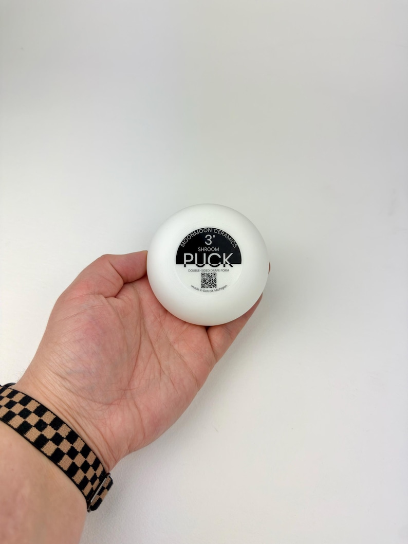 May include: A white, circular ceramic tool labeled Moonmoon Ceramics 3" Shroom Puck, Double-Sided Drape Form, with a QR code and text indicating it was made in Detroit, Michigan. The object is smooth, round, and features black printed text.