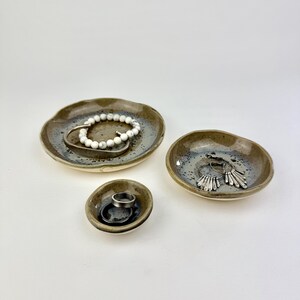 May include: Three small, round, handmade ceramic dishes with irregular, wavy edges. The dishes feature a speckled, mottled glaze in shades of brown, gray, and cream. They contain various pieces of jewelry, including a white beaded bracelet and rings.