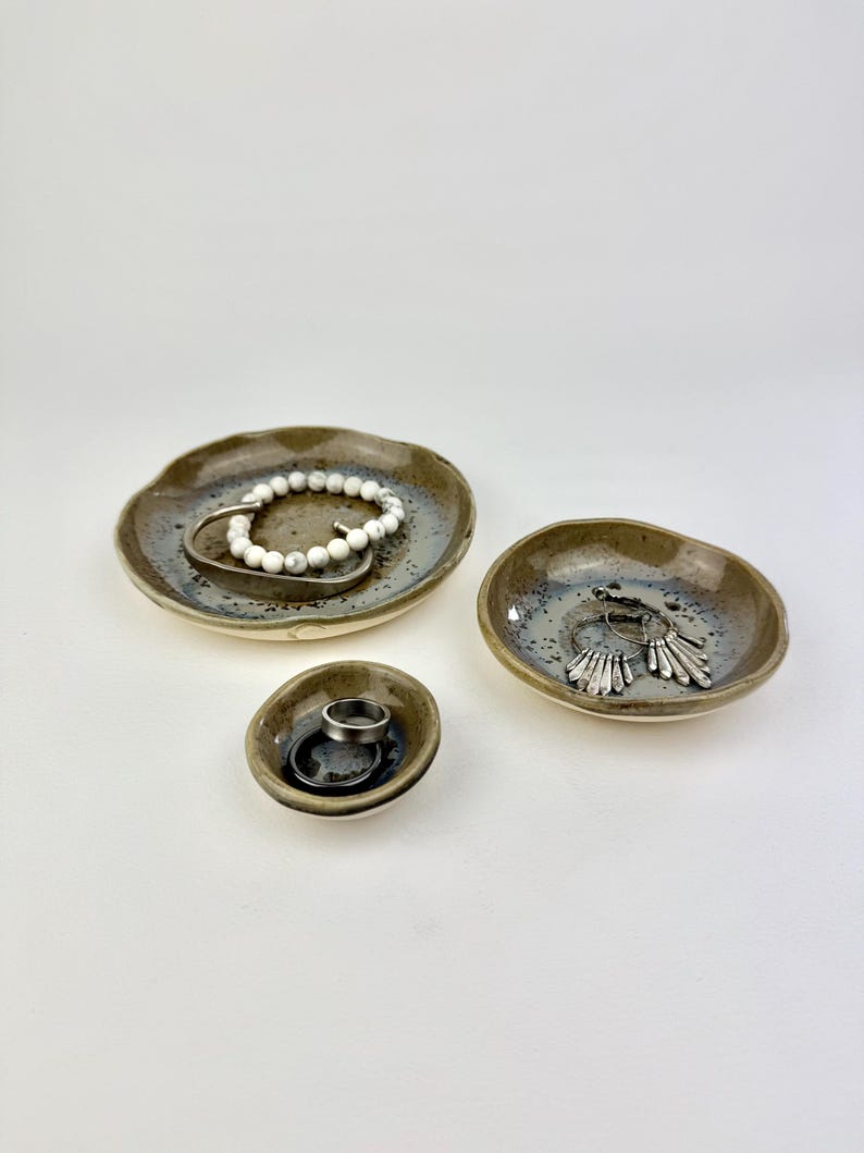 May include: Three small, round, handmade ceramic dishes with irregular, wavy edges. The dishes feature a speckled, mottled glaze in shades of brown, gray, and cream. They contain various pieces of jewelry, including a white beaded bracelet and rings.