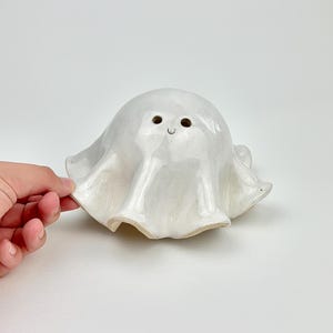 Op de afbeelding: Een handgemaakte, witte keramische spookfiguur met een glanzende afwerking. Het spook heeft een afgerond lichaam en een gedrapeerde, vloeiende vorm. Het heeft twee kleine oogjes en een gebogen mond, waardoor het een vriendelijke uitstraling heeft.