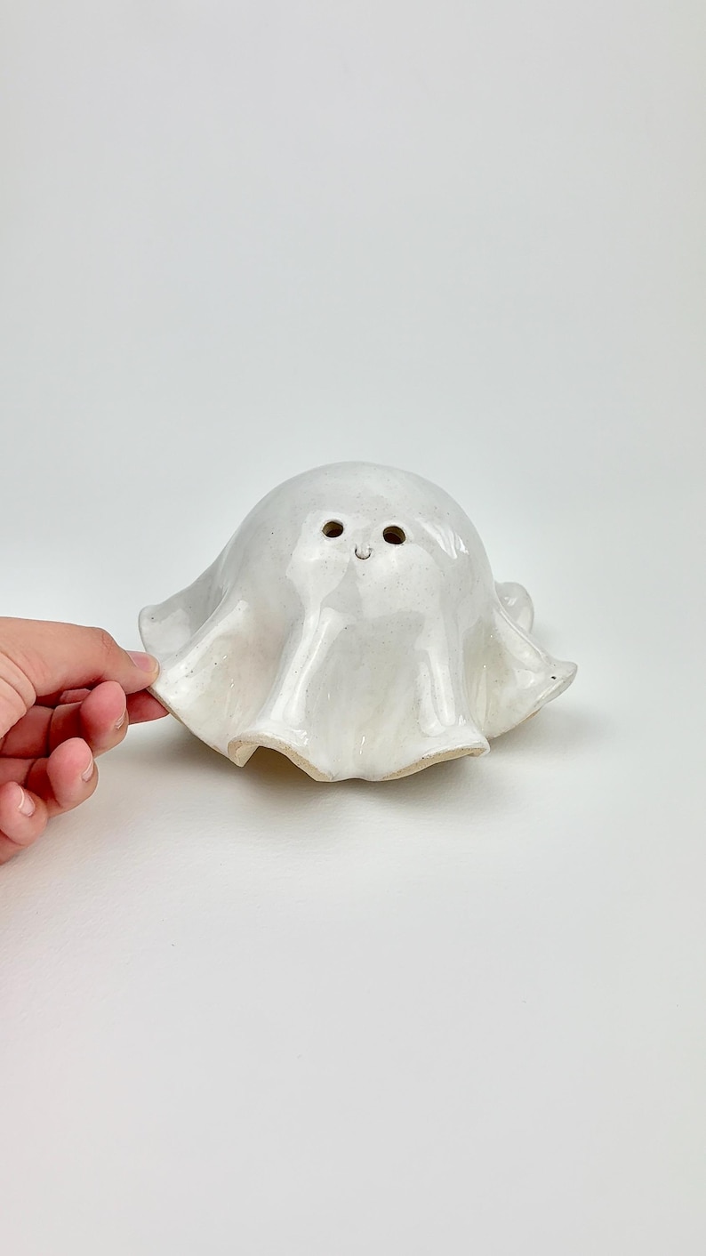 Op de afbeelding: Een handgemaakte, witte keramische spookfiguur met een glanzende afwerking. Het spook heeft een afgerond lichaam en een gedrapeerde, vloeiende vorm. Het heeft twee kleine oogjes en een gebogen mond, waardoor het een vriendelijke uitstraling heeft.