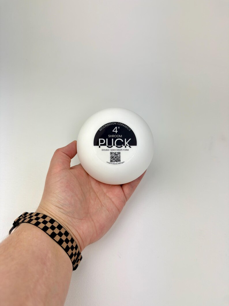 May include: A white, circular ceramic drape form with a black label in the center. The label reads MOONMOON CERAMICS 4" SHROOM PUCK DOUBLE-SIDED DRAPE FORM and includes a QR code. The object is a smooth, solid, rounded tool for pottery shaping.