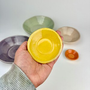 May include: A collection of five small ceramic bowls in various colors including bright yellow, sage green, muted purple, beige, and a tiny orange bowl. Each bowl features a smooth, glossy finish with subtle speckling throughout the glaze.
