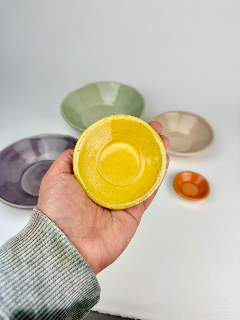 May include: A collection of five small ceramic bowls in various colors including bright yellow, sage green, muted purple, beige, and a tiny orange bowl. Each bowl features a smooth, glossy finish with subtle speckling throughout the glaze.