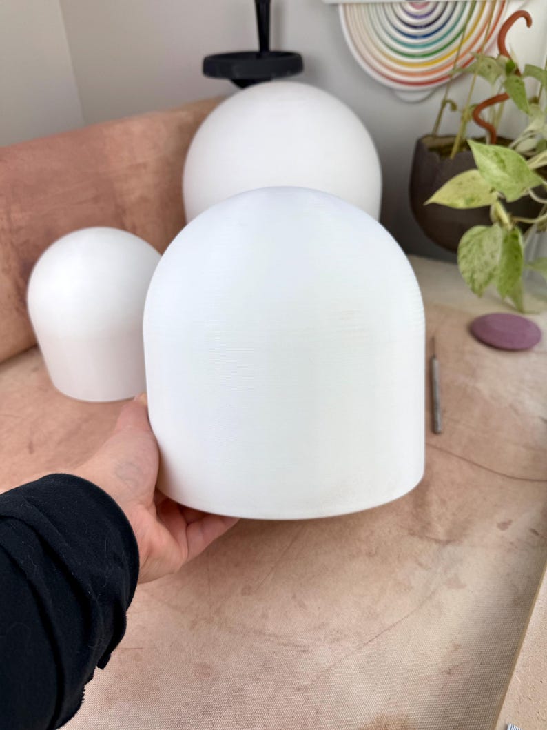 May include: Three smooth, matte white, dome-shaped objects with a cylindrical base. The objects are solid in color and feature a rounded top. One dome is held in the foreground, while two additional domes of varying sizes are positioned behind it.
