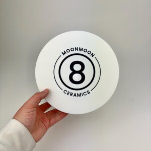 May include: A circular white ceramic object featuring a black graphic design in the center. The design consists of a large number 8 enclosed within two concentric circles, with the text MOONMOON CERAMICS printed in a circular arc around the logo.