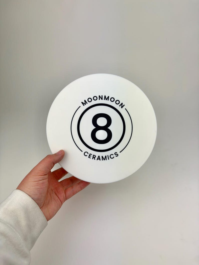 May include: A circular white ceramic object featuring a black graphic design in the center. The design consists of a large number 8 enclosed within two concentric circles, with the text MOONMOON CERAMICS printed in a circular arc around the logo.