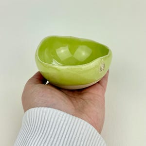 Op de afbeelding: Een kleine, handgemaakte keramische kom met een levendig, glanzend groen interieur en een lichtgroen exterieur. De kom heeft een organische, onregelmatige vorm en een witte basis. Een kleine, rode stempel is zichtbaar aan de zijkant.