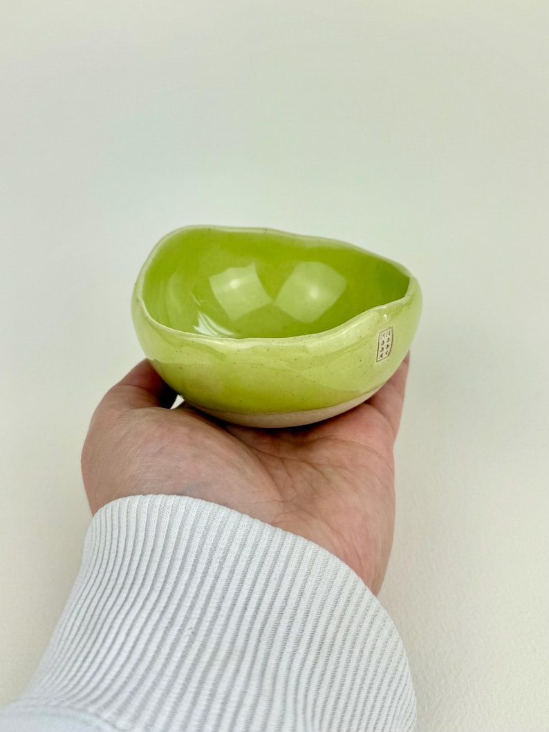 Op de afbeelding: Een kleine, handgemaakte keramische kom met een levendig, glanzend groen interieur en een lichtgroen exterieur. De kom heeft een organische, onregelmatige vorm en een witte basis. Een kleine, rode stempel is zichtbaar aan de zijkant.