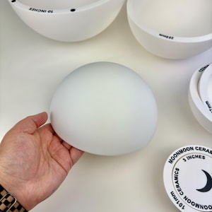 May include: A collection of white ceramic bowls and a circular lid, featuring black printed text including Moonmoon Ceramics and measurements in inches. One bowl is held, showing a smooth, matte, hemispherical shape with a clean white finish.