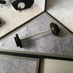 May include: A miniature barbell with black weights and a silver bar is positioned in the center of a diorama. The scene includes a small gym setting with black exercise equipment and a white brick wall.