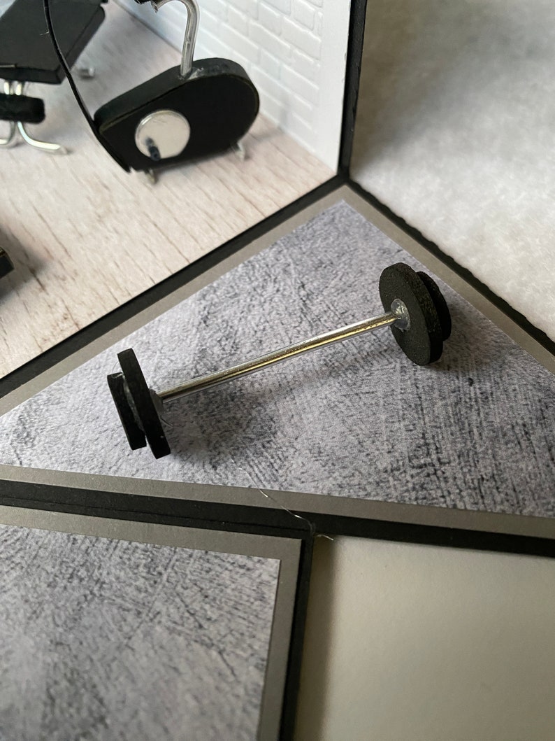May include: A miniature barbell with black weights and a silver bar is positioned in the center of a diorama. The scene includes a small gym setting with black exercise equipment and a white brick wall.