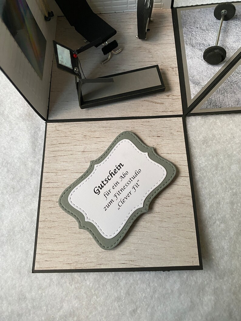 May include: A handmade pop-up card with a gym theme. The card includes miniature gym equipment such as a weight bench, treadmill, and barbell. A decorative label reads "Gutschein f&uuml;r ein Abo zum Fitnessstudio 'Clever Fit'" in black text.