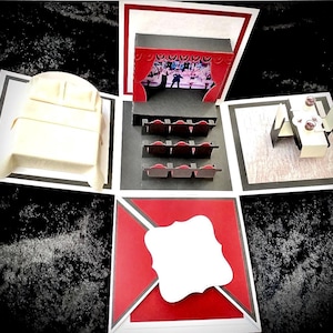 Könnte beinhalten: Eine handgefertigte Papierkunstbox mit mehreren Ebenen. Die Box öffnet sich und zeigt eine Theaterbühne mit rotem Vorhang, Sitzplätzen, einem Bett und einem Esstisch. Die Box ist weiß, rot und schwarz, mit einem leeren weißen Etikett.