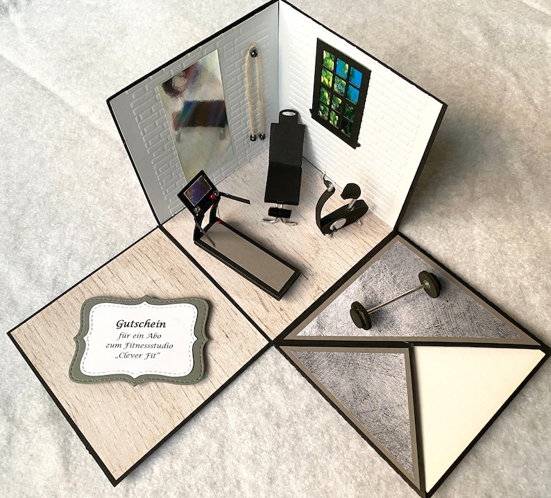 May include: A handmade papercraft diorama of a gym, featuring a treadmill, weight bench, and exercise bike. The scene includes a barbell and a gift certificate with text. The walls are white with a brick pattern, and the floor is light wood.
