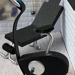 May include: Miniature black exercise bike with silver detailing. The bike features a black seat and a silver handlebar with a blue gem. The bike is positioned against a white brick wall.