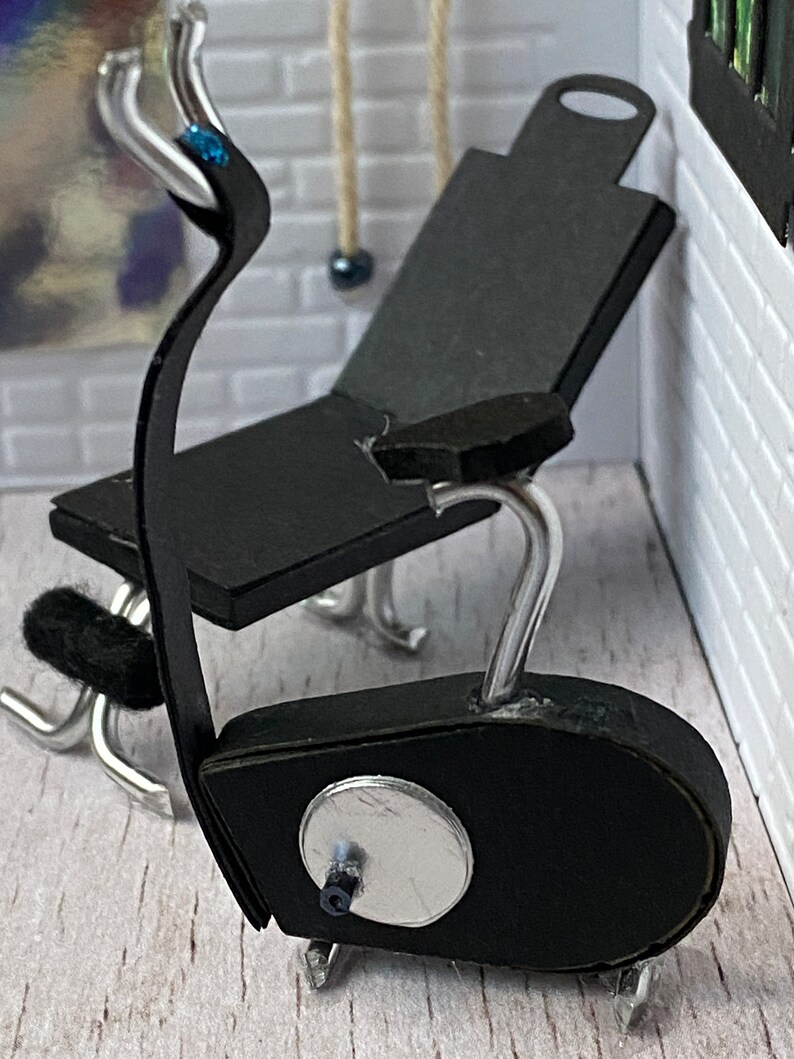 May include: Miniature black exercise bike with silver detailing. The bike features a black seat and a silver handlebar with a blue gem. The bike is positioned against a white brick wall.