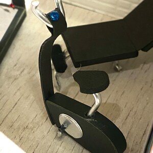 May include: A small black chair with silver metal detailing and a blue gemstone. The chair features a curved back and a circular base. It is set on a light-coloured surface.