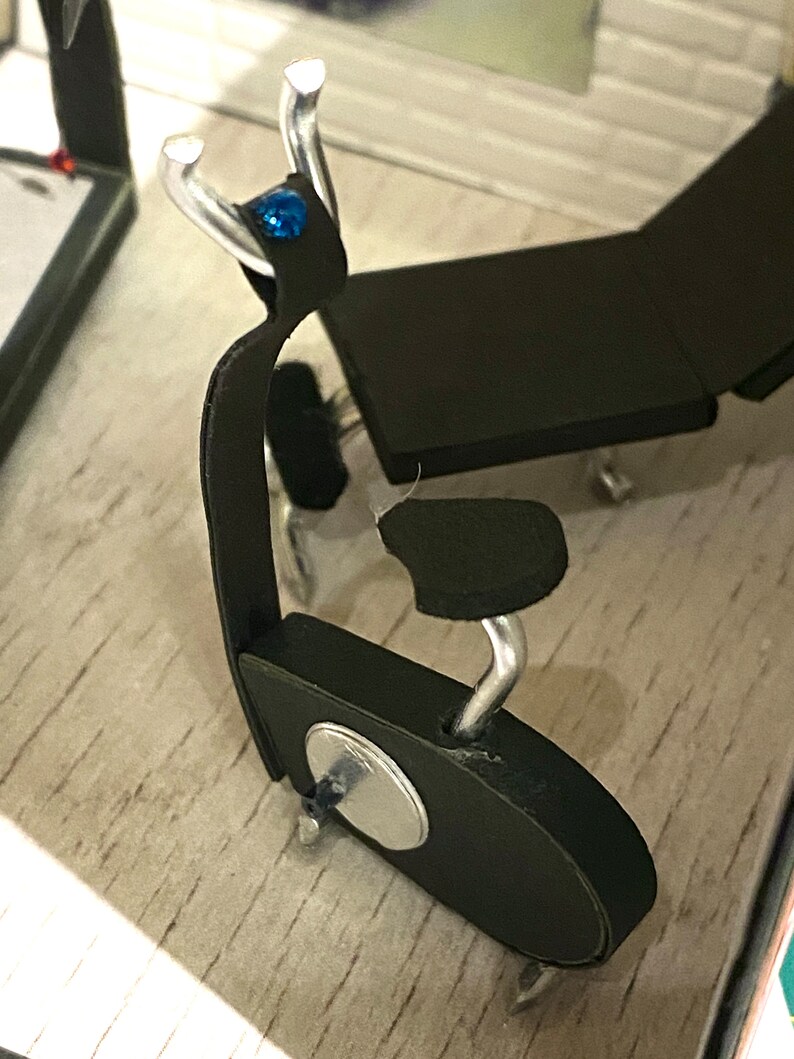 May include: A small black chair with silver metal detailing and a blue gemstone. The chair features a curved back and a circular base. It is set on a light-coloured surface.