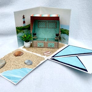 Op de afbeelding: Een handgemaakte pop-upkaart met een strandscène. De kaart opent om een miniatuur strandcabana te onthullen met een vuurtorenachtergrond, schelpen en een kleine potplant. De kaart heeft een blauw en wit geometrisch ontwerp.