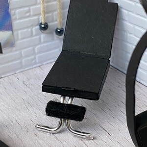 May include: A miniature black chair with a rectangular seat and back, and a small handle at the top. The chair has a silver metal base and a black fuzzy band. The chair is set against a white brick wall.