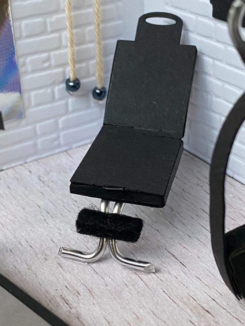 May include: A miniature black chair with a rectangular seat and back, and a small handle at the top. The chair has a silver metal base and a black fuzzy band. The chair is set against a white brick wall.