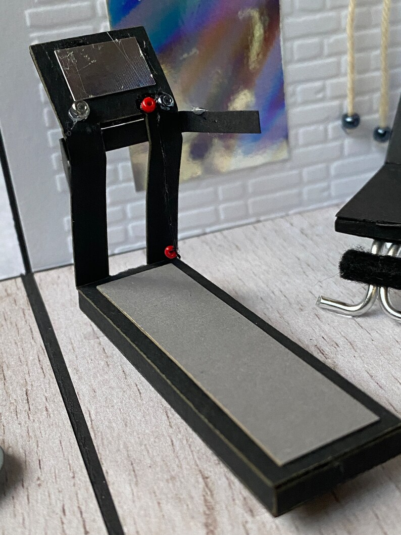 May include: A miniature black treadmill with a grey running surface and a silver display. The treadmill has red and clear accents, set against a white brick wall. A small black exercise bench is visible.